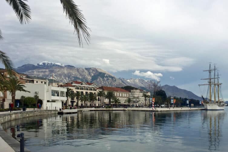 Panoramic view of the Tivat waterfront on the Bay of Kotor, featuring a long promenade lined with palm trees and modern white buildings, with the historic sailing ship 'Jadran' docked on the right and snow-capped mountains in the distance.