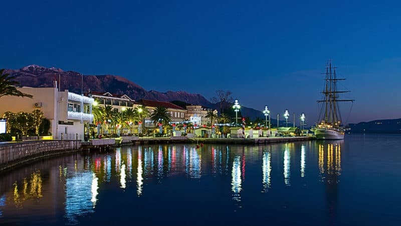 Night view of the brightly lit promenade and waterfront buildings in Tivat, Montenegro, with the historic sailing ship 'Jadran' docked on the right and mountain silhouettes reflected on the calm water of the Bay of Kotor.
