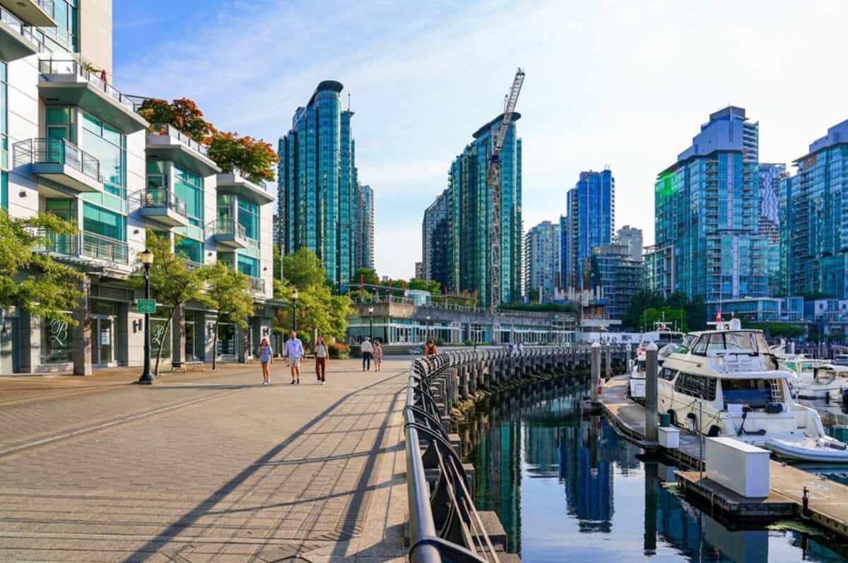 Scenic waterfront with docked boats, glass high-rise buildings, and tree-lined pedestrian walkway in urban Vancouver, BC, Canada