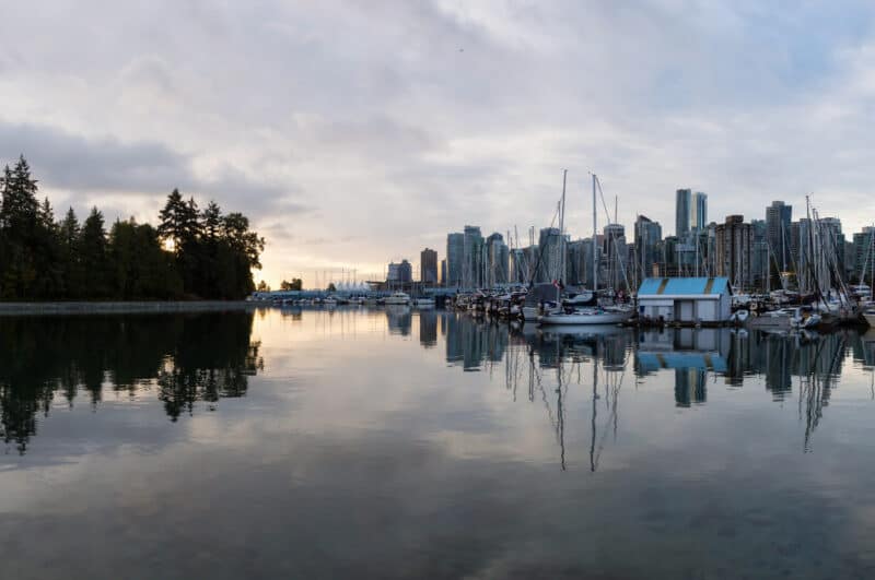 Peaceful marina with sailboats, city skyline, and forested shoreline reflecting on calm water under overcast sky