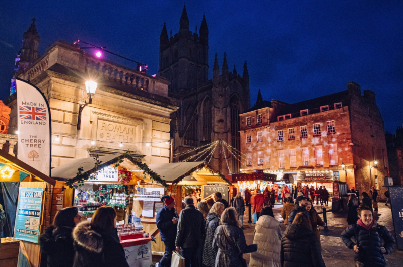 Wooden chalets and shoppers at the Bath Christmas Market outside Roman Baths and Abbey at dusk.