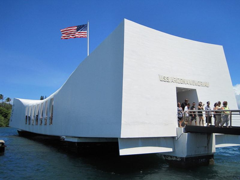 American flag flies atop the USS Arizona Memorial at Pearl Harbor. At right, tourists exit the building.