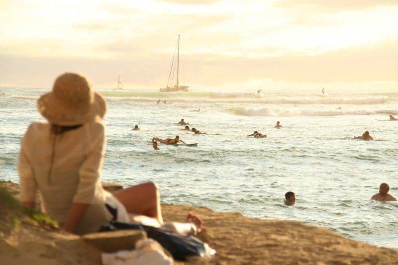 Woman wearing a hat seated on a beach. Looking at people swimming in the water. Sailboat in the background.